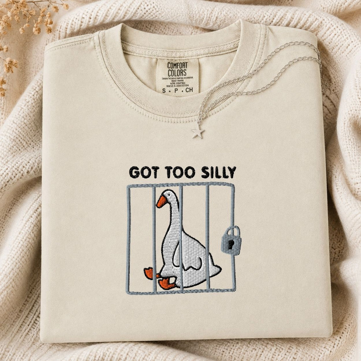 A folded cream-colored tshirt with a graphic of a goose in a cage and the text 'GOT TOO SILLY' on the front, styled on a beige knit blanket.