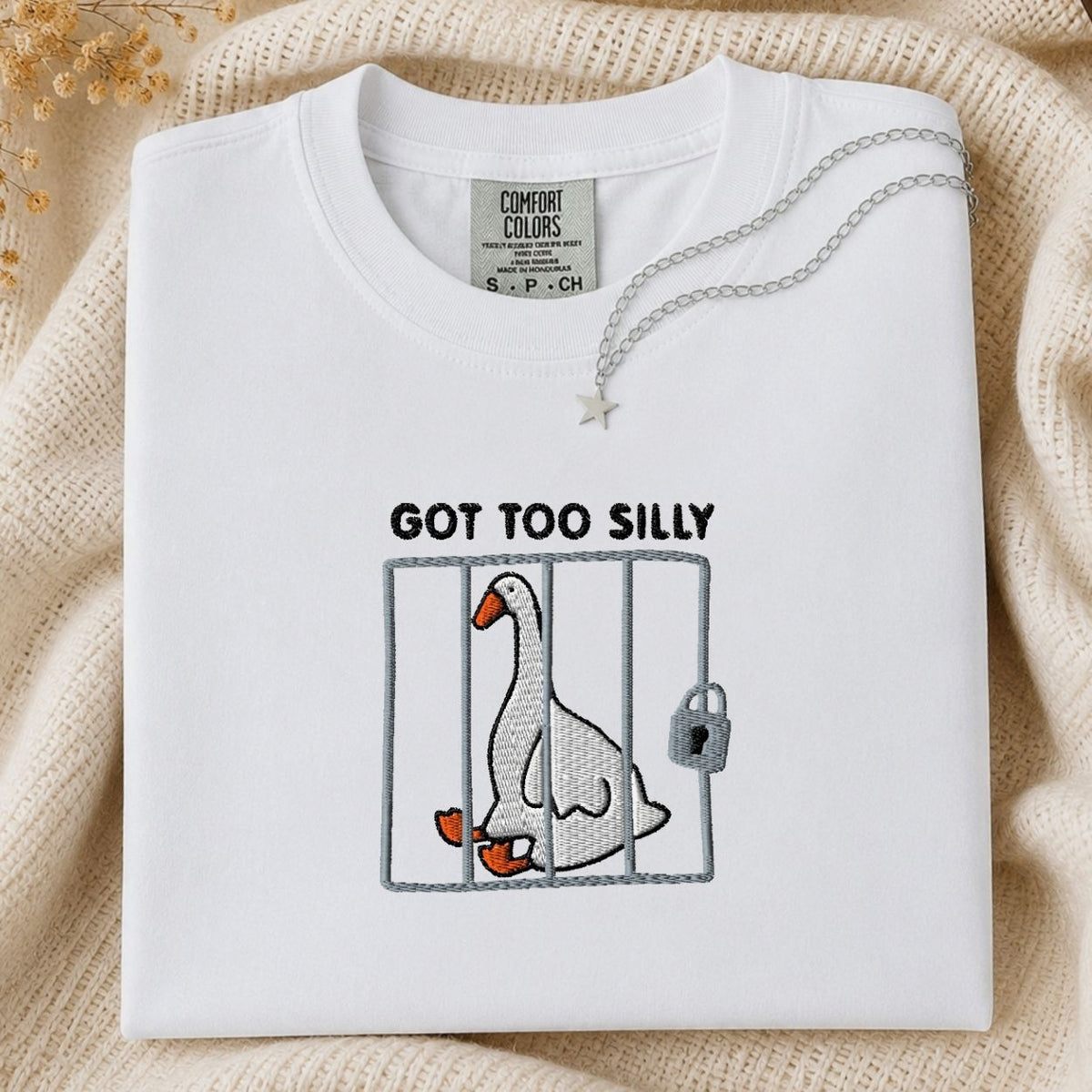 A folded white Comfort Colors t-shirt with a graphic of a goose in a cage and the text 'GOT TOO SILLY'. The shirt is styled on a beige knit blanket with a silver necklace draped over the collar.