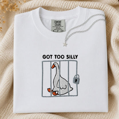 A folded white Comfort Colors t-shirt with a graphic of a goose in a cage and the text 'GOT TOO SILLY'. The shirt is styled on a beige knit blanket with a silver necklace draped over the collar.