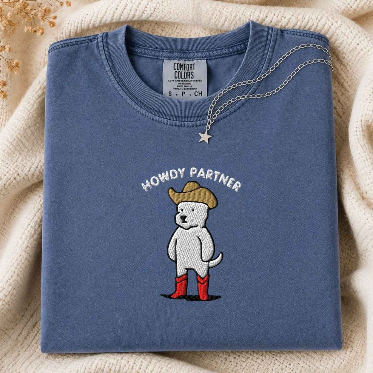 A folded slate blue t-shirt with a graphic of a white dog wearing a cowboy hat and red boots, with the text 'HOWDY PARTNER' above it. The shirt is styled on a cream-colored knit blanket with a small silver star necklace draped over the collar.