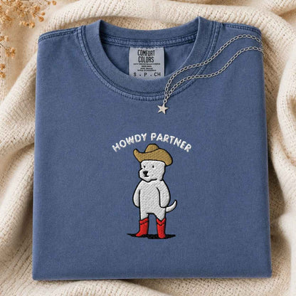 A folded slate blue t-shirt with a graphic of a white dog wearing a cowboy hat and red boots, with the text 'HOWDY PARTNER' above it. The shirt is styled on a cream-colored knit blanket with a small silver star necklace draped over the collar.
