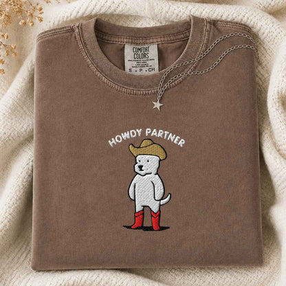 A folded brown tshirt with a graphic of a white dog wearing a cowboy hat and red boots, under the text 'HOWDY PARTNER'. A silver star necklace is draped over the collar.
