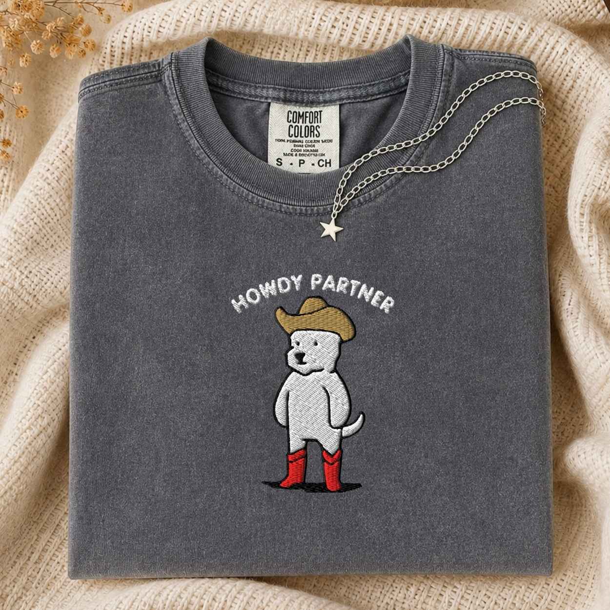 A folded dark gray t-shirt with a western-themed embroidered dog graphic and the text 'HOWDY PARTNER'. The shirt is styled on a beige knit blanket with a silver star necklace draped over the collar.