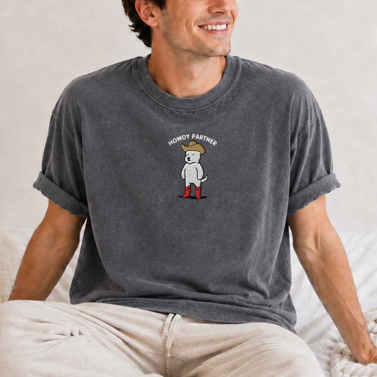 A man wearing a dark gray heathered t-shirt with a cartoon graphic of a white dog wearing a tan hat and red boots, with the text 'HONORY PARTNER' above it. The man is sitting on a bed with light-colored sheets.