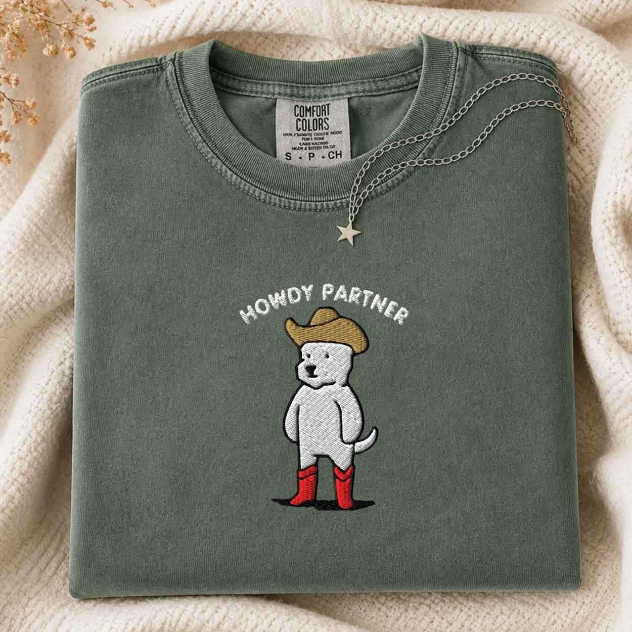 A folded sage green Comfort Colors t-shirt featuring a cartoon dog wearing a cowboy hat, red boots, and the text 'HOWDY PARTNER' embroidered on the front. The shirt is styled on a soft cream knit blanket with a silver star necklace draped over it.