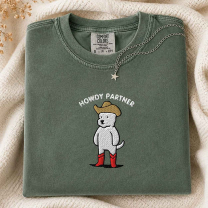 A folded sage green Comfort Colors t-shirt featuring a cartoon dog wearing a cowboy hat, red boots, and the text 'HOWDY PARTNER' embroidered on the front. The shirt is styled on a soft cream knit blanket with a silver star necklace draped over it.