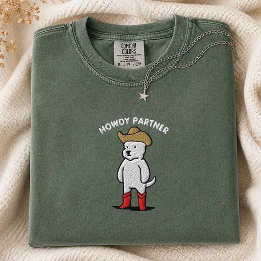 A folded sage green Comfort Colors t-shirt featuring a cartoon dog wearing a cowboy hat, red boots, and the text 'HOWDY PARTNER' embroidered on the front. The shirt is styled on a soft cream knit blanket with a silver star necklace draped over it.