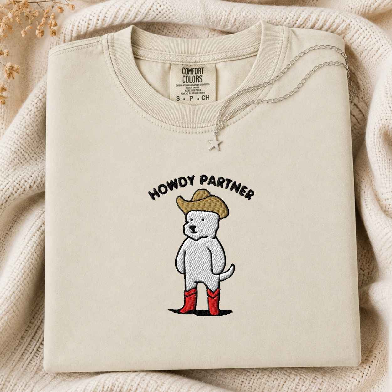 A folded cream-colored tshirt with a graphic of a white dog wearing a tan cowboy hat, red boots, and the text 'HOWDY PARTNER' on the front. The sweatshirt is displayed on a cozy, textured cream knit blanket with a small silver star pendant resting on the chest.