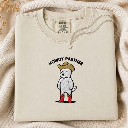 A folded cream-colored tshirt with a graphic of a white dog wearing a tan cowboy hat, red boots, and the text 'HOWDY PARTNER' on the front. The sweatshirt is displayed on a cozy, textured cream knit blanket with a small silver star pendant resting on the chest.