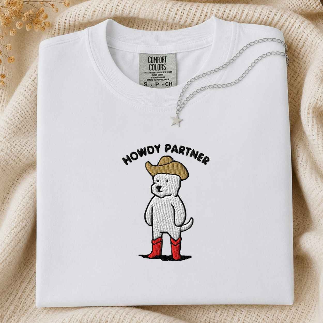 A folded white Comfort Colors t-shirt with a 'HOWDY PARTNER' graphic of a cartoon dog in a cowboy hat and red boots, styled on a beige knit blanket with a silver necklace.