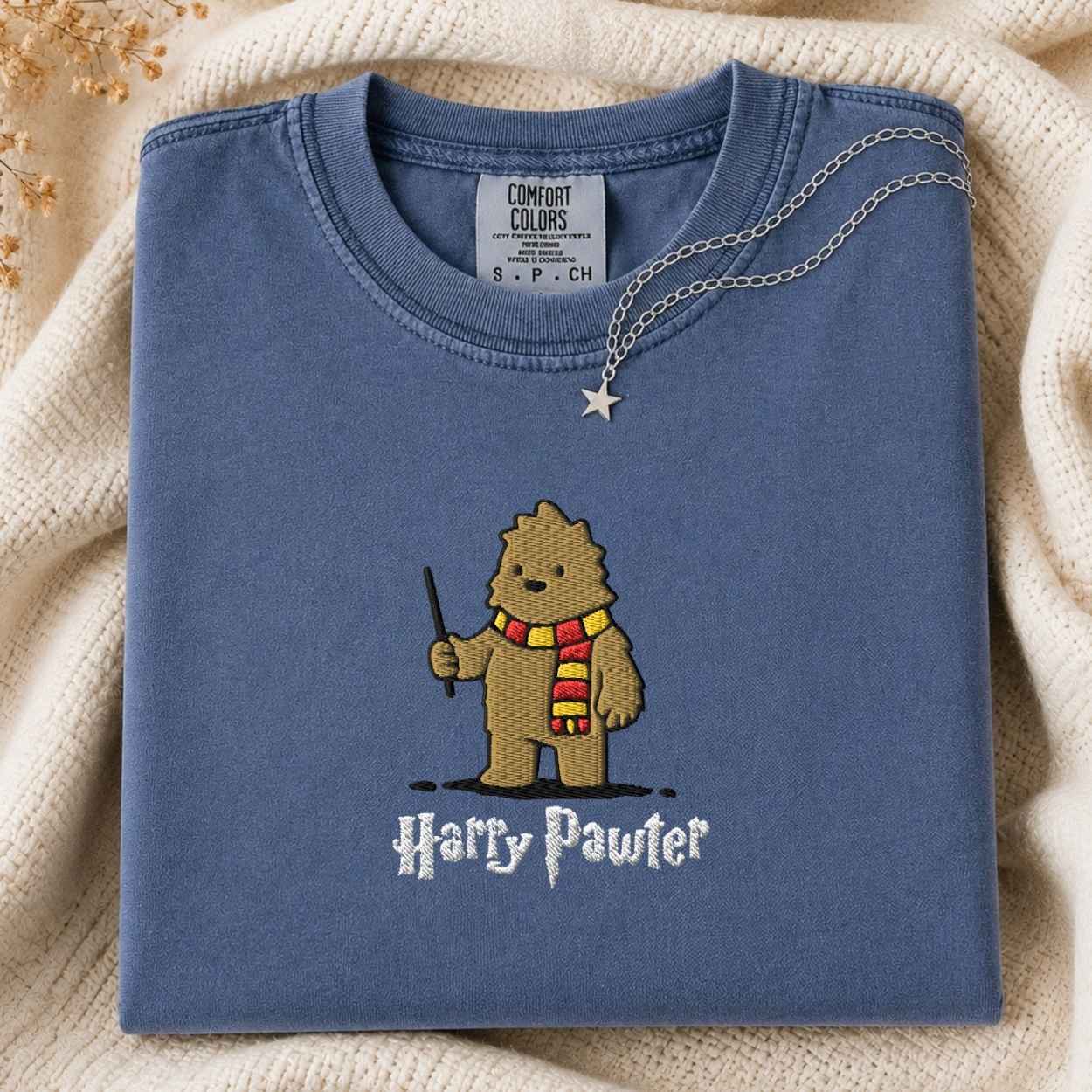 A folded slate blue t-shirt with a cartoon Bigfoot character wearing a red and yellow striped scarf and holding a wand, with the text 'Harry Pawter' below. A silver necklace with a star pendant is draped over the collar.