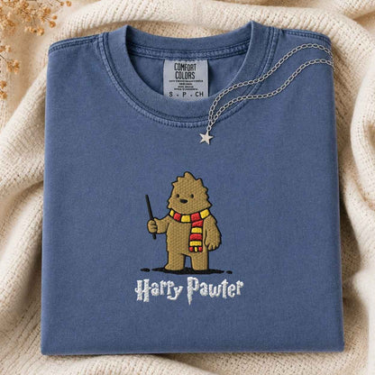 A folded slate blue t-shirt with a cartoon Bigfoot character wearing a red and yellow striped scarf and holding a wand, with the text 'Harry Pawter' below. A silver necklace with a star pendant is draped over the collar.