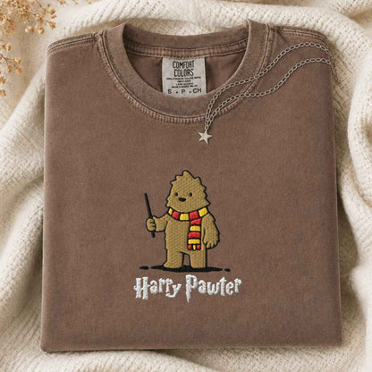 A folded sage brown Comfort Colors t-shirt featuring an art style embroidered bigfoot with a wand and a red and yellow striped scarf, with the text 'Harry Pawter' embroidered below. The shirt is styled on a cream knit blanket with a silver necklace draped over the collar.
