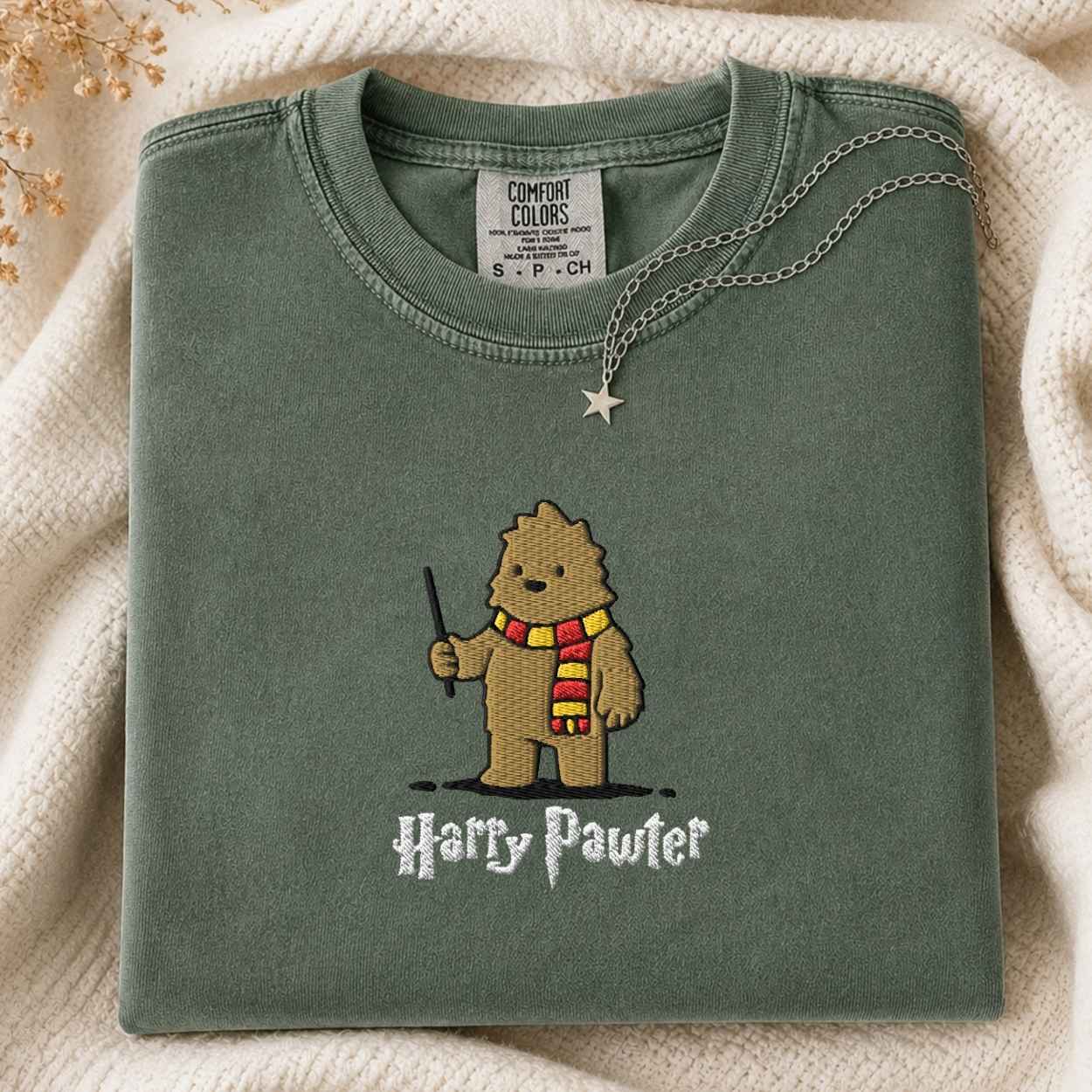 A folded sage green Comfort Colors t-shirt featuring an art style embroidered bigfoot with a wand and a red and yellow striped scarf, with the text 'Harry Pawter' embroidered below. The shirt is styled on a cream knit blanket with a silver necklace draped over the collar.