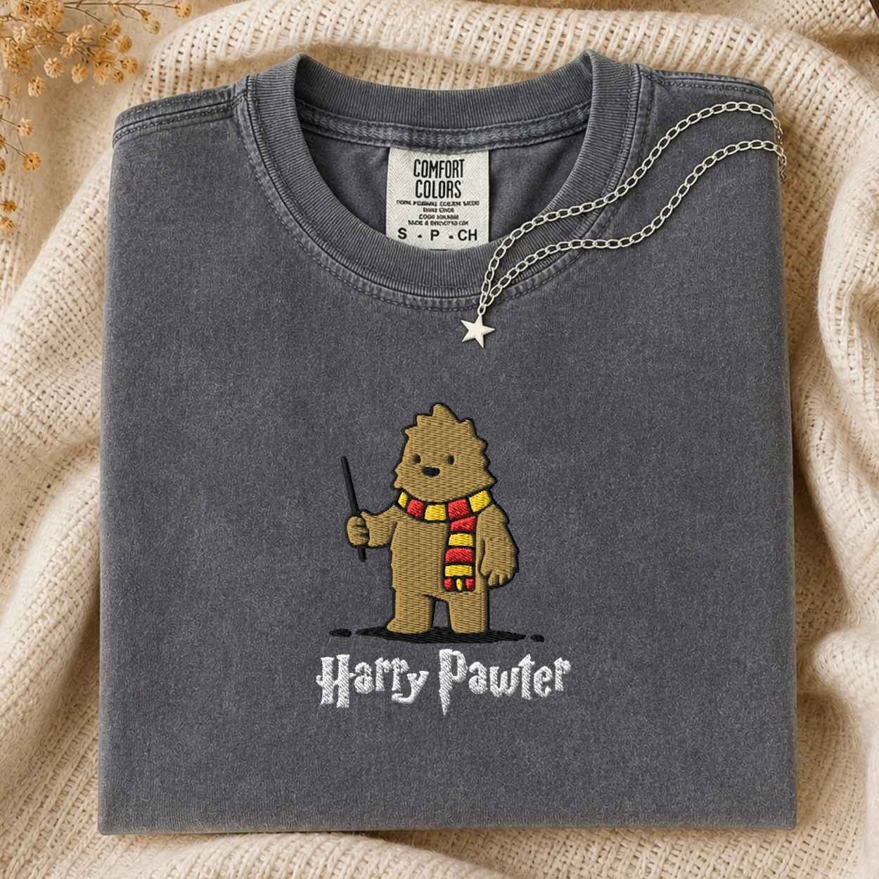 A folded sage gray Comfort Colors t-shirt featuring an art style embroidered bigfoot with a wand and a red and yellow striped scarf, with the text 'Harry Pawter' embroidered below. The shirt is styled on a cream knit blanket with a silver necklace draped over the collar.
