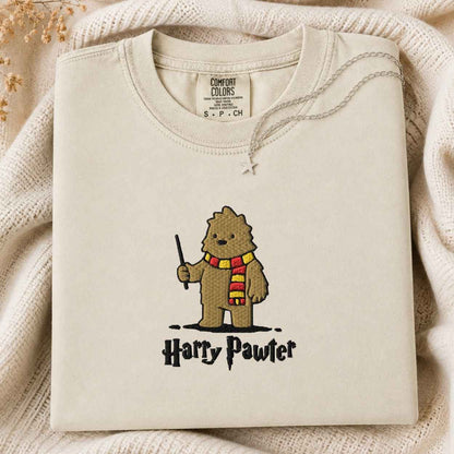 A folded sage ivory Comfort Colors t-shirt featuring an art style embroidered bigfoot with a wand and a red and yellow striped scarf, with the text 'Harry Pawter' embroidered below. The shirt is styled on a cream knit blanket with a silver necklace draped over the collar.
