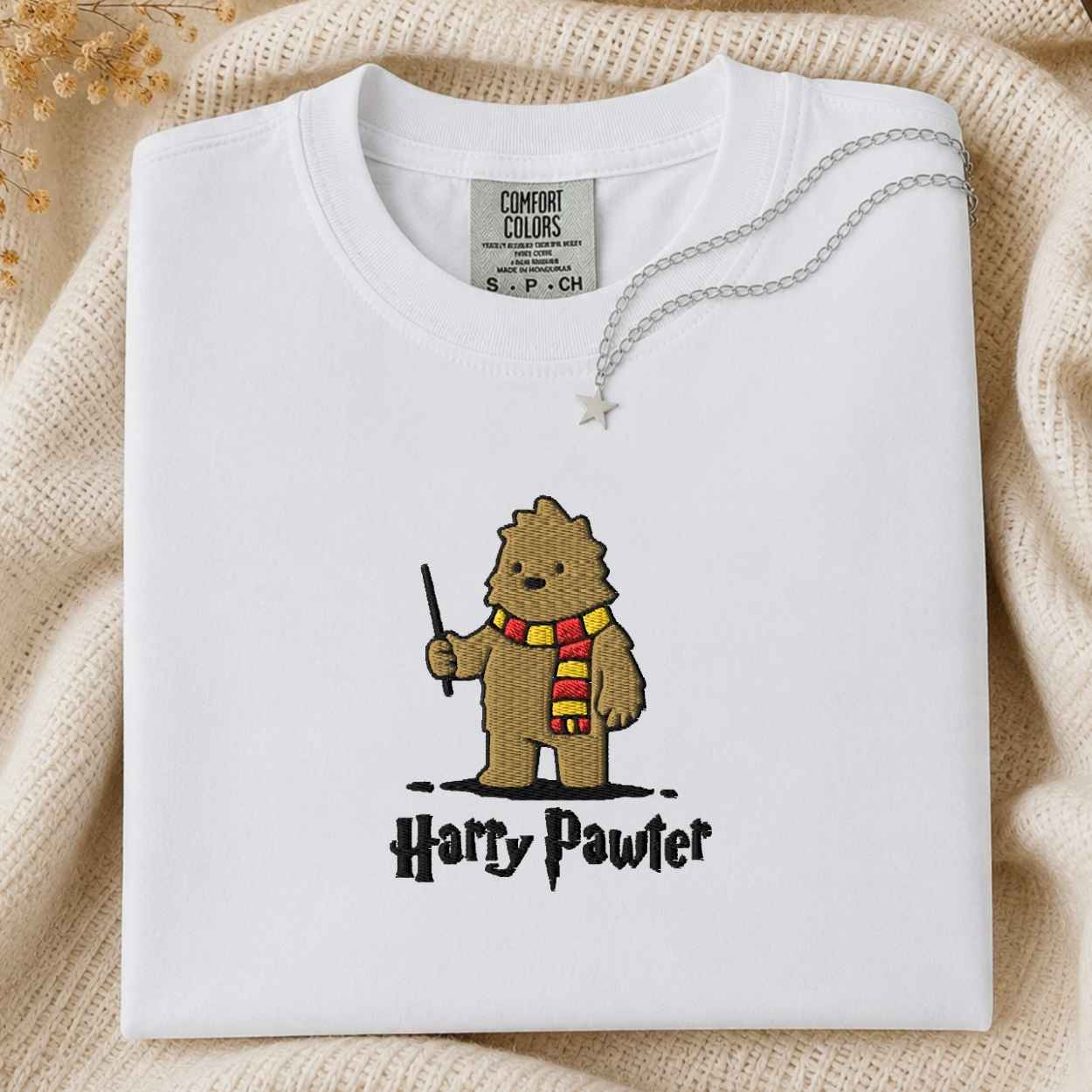 A folded white Comfort Colors t-shirt featuring a cartoon graphic of a bigfoot wearing a red and yellow striped scarf and holding a wand, with the text 'Harry Pawter' underneath. The shirt is styled on a beige knit blanket with a silver necklace draped over the collar.