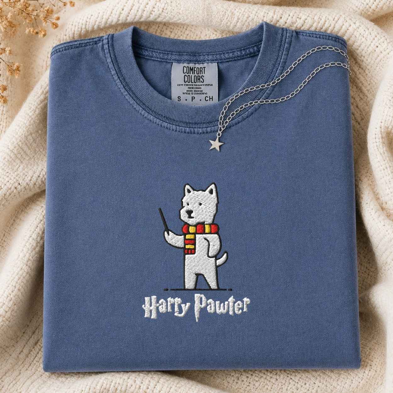 A folded dusty blue t-shirt with an embroidered white dog character wearing a red and yellow striped scarf and holding a wand, with the text 'Harry Pawter' embroidered below. A silver necklace with a star pendant is draped over the collar.