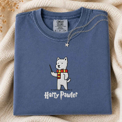 A folded dusty blue t-shirt with an embroidered white dog character wearing a red and yellow striped scarf and holding a wand, with the text 'Harry Pawter' embroidered below. A silver necklace with a star pendant is draped over the collar.