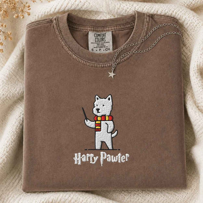 A folded brown T-shirt with a printed cartoon dog wearing a red and yellow striped scarf and holding a wand, with the text 'Harry Pawter' below. The shirt is styled on a cream knit blanket with a silver star pendant draped over the collar.