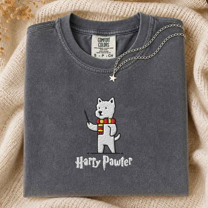 A folded dark gray t-shirt with a white embroidered dog character wearing a red and yellow striped scarf, with the text 'Harry Pawter' below. The shirt is styled on a beige knit blanket with a silver necklace draped over the collar.