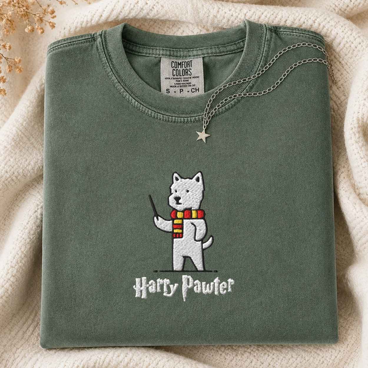 A folded sage green t-shirt featuring an embroidered white dog character wearing a red and yellow striped scarf and holding a wand, with 'Harry Pawter' embroidered below. The shirt is styled on a cream knit blanket with a silver star necklace draped over the collar.