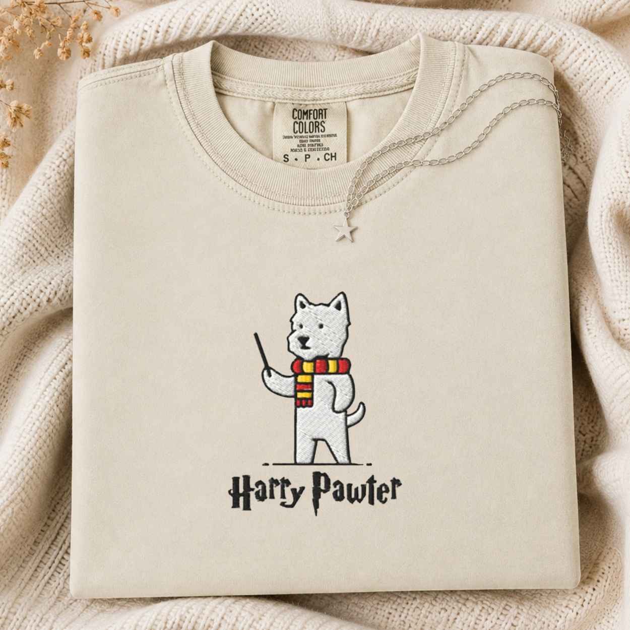 A folded cream-colored t-shirt with a graphic of a white dog wearing a red and yellow striped scarf and holding a wand, with the text 'Harry Pawter' underneath. The shirt is displayed on a cozy, textured cream knit blanket with dried flowers in the corner.