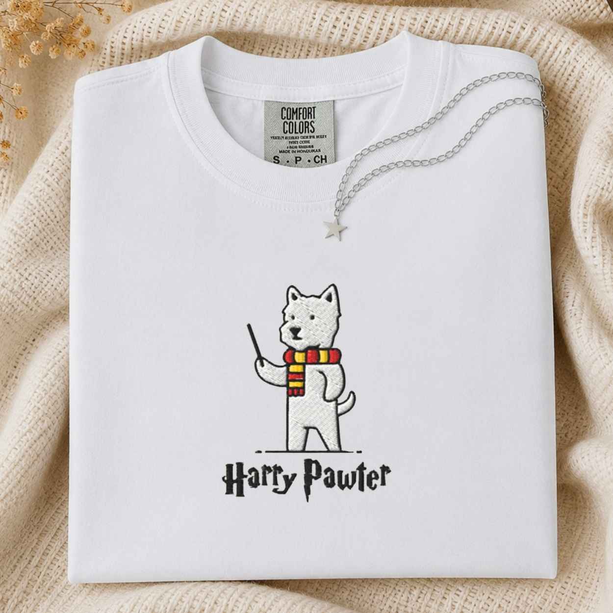 A folded white Comfort Colors T-shirt with a printed illustration of a white dog wearing a red and yellow striped scarf and holding a wand, with the text 'Harry Pawter' below. The shirt is styled on a beige knit blanket with a silver necklace draped across the collar.