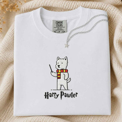 A folded white Comfort Colors T-shirt with a printed illustration of a white dog wearing a red and yellow striped scarf and holding a wand, with the text 'Harry Pawter' below. The shirt is styled on a beige knit blanket with a silver necklace draped across the collar.