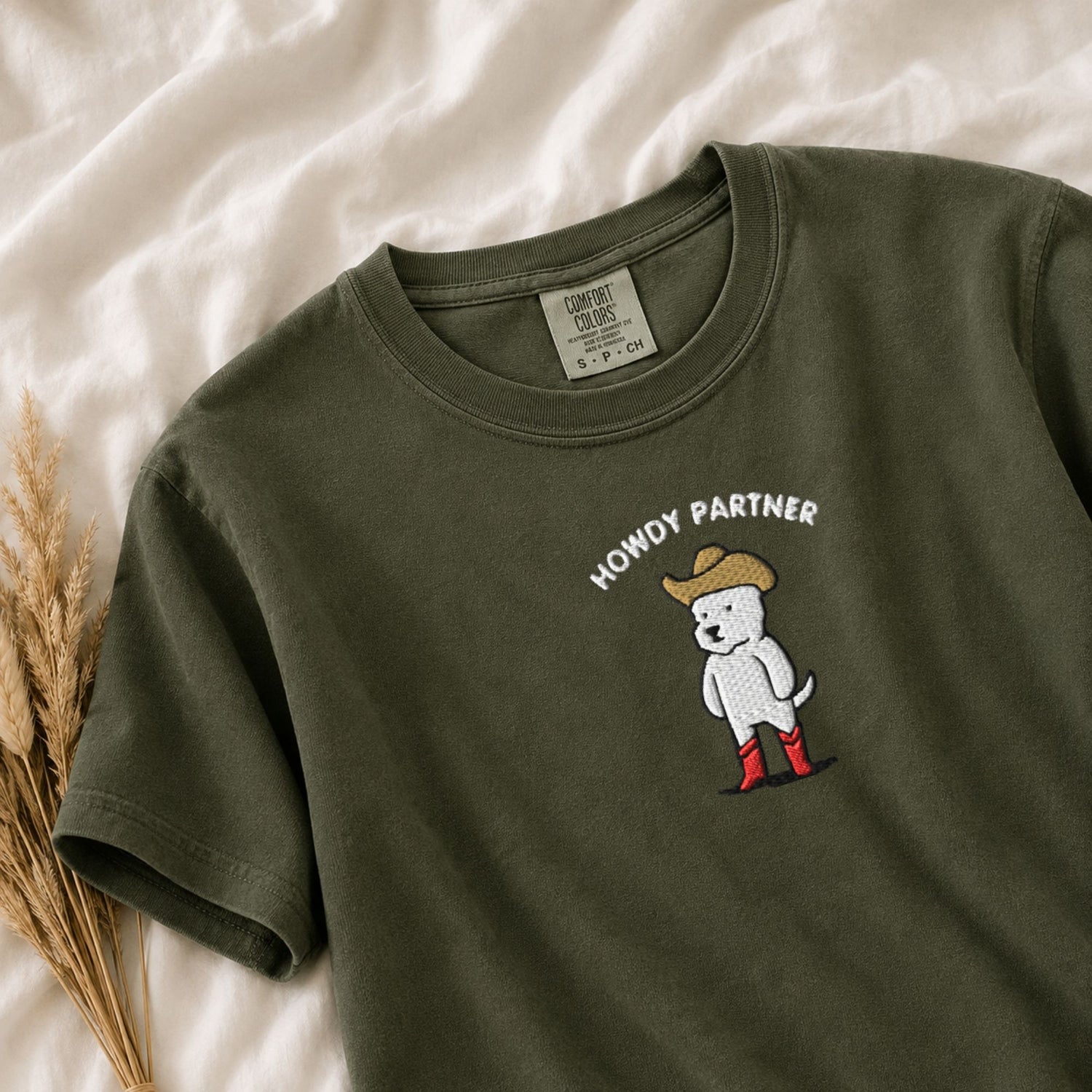 An olive green short-sleeve t-shirt laid flat on a white fabric background, featuring a white embroidered dog wearing a tan cowboy hat and red boots with the text 'HOWDY PARTNER' above it. A small woven brand label is visible inside the collar, and dried wheat stems decorate the lower left corner.