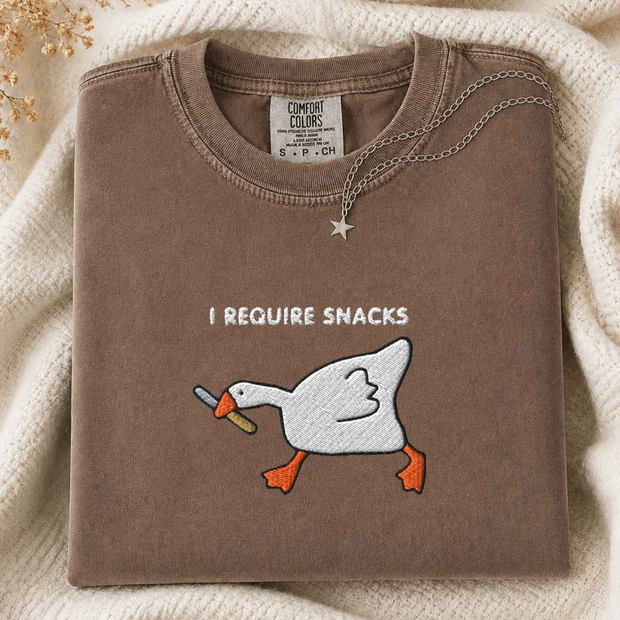 A folded brown crewneck tshirt with a embroidery graphic of a white goose with a knife and the text 'I REQUIRE SNACKS' on the front. The sweatshirt is styled on a cream knit blanket with a silver star necklace draped over the collar.