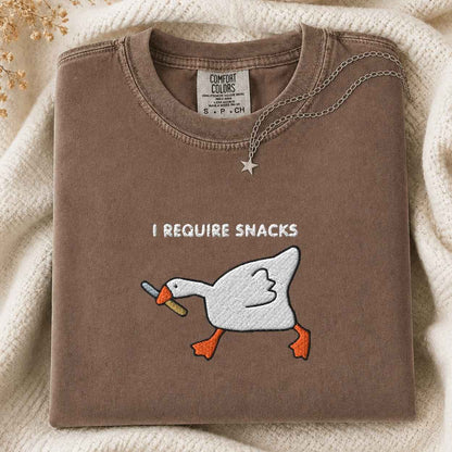 A folded brown crewneck tshirt with a embroidery graphic of a white goose with a knife and the text 'I REQUIRE SNACKS' on the front. The sweatshirt is styled on a cream knit blanket with a silver star necklace draped over the collar.