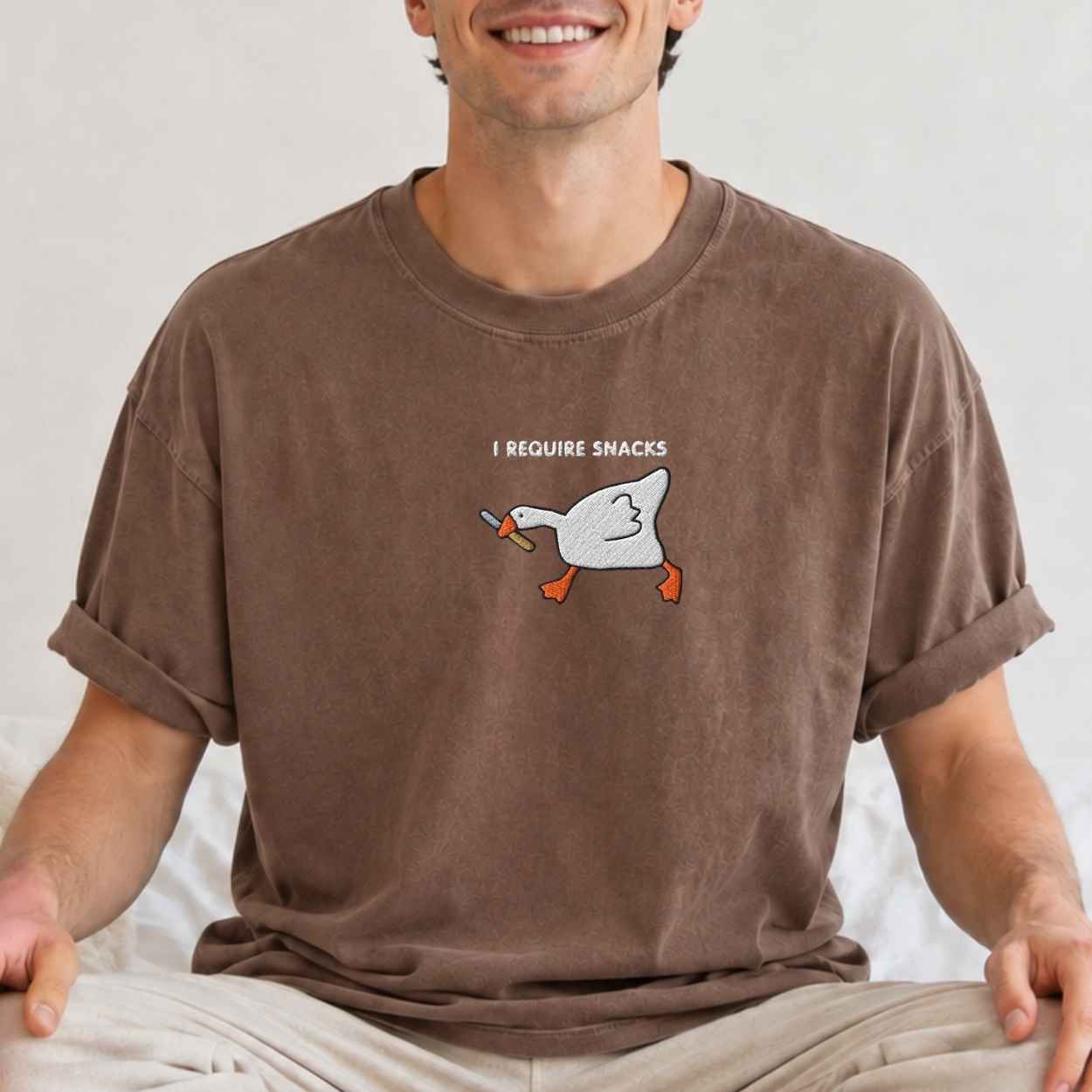 A man wearing a brown crewneck t-shirt with an embroidery graphic of a white goose holding a knife and the text 'I REQUIRE SNACKS' on the chest.