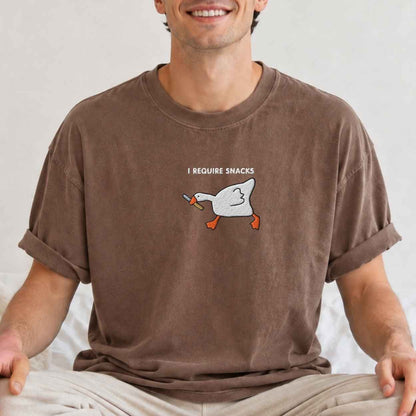 A man wearing a brown crewneck t-shirt with an embroidery graphic of a white goose holding a knife and the text 'I REQUIRE SNACKS' on the chest.