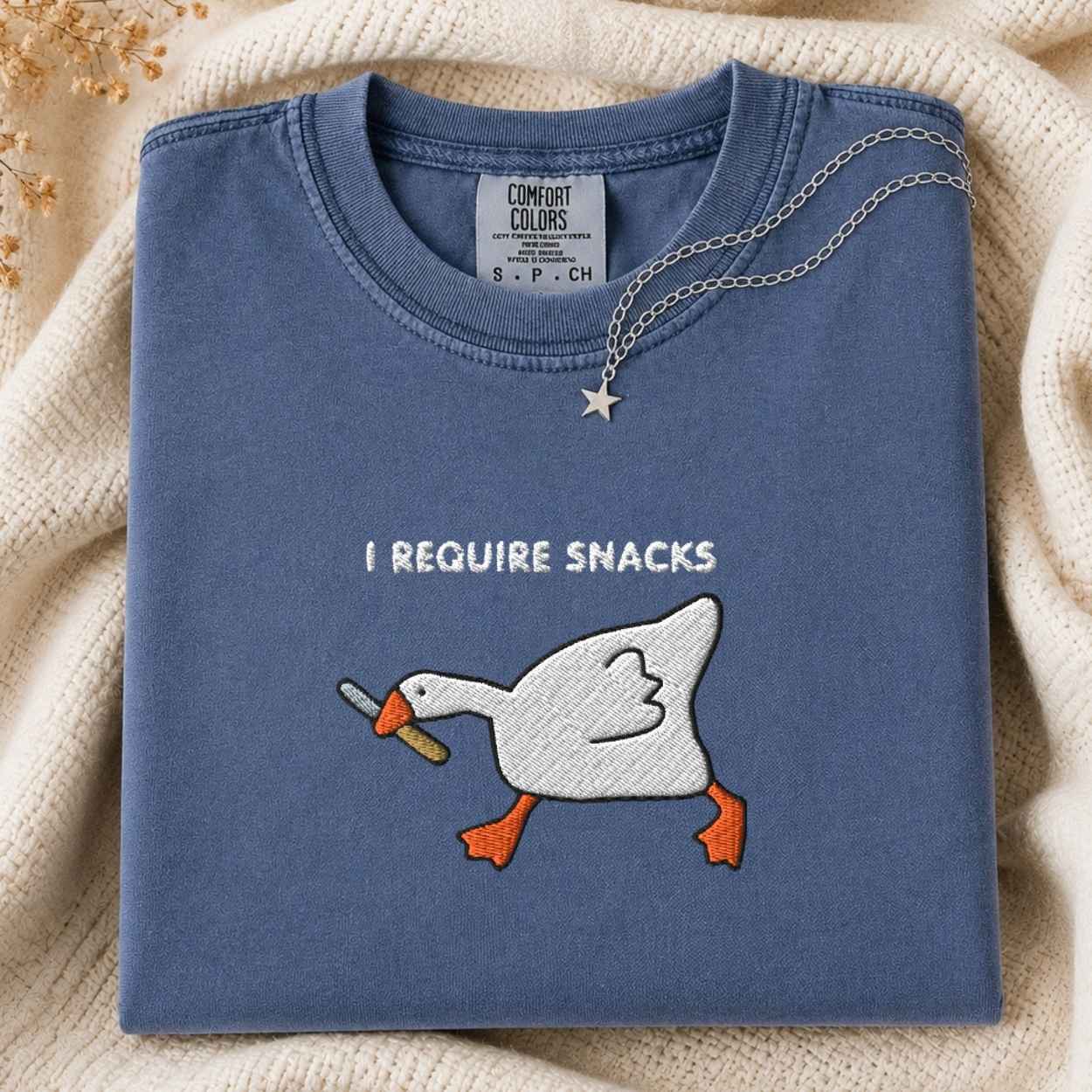 A folded slate blue crewneck tshirt with a white goose embroidery graphic holding a knife and the text 'I REQUIRE SNACKS'. A silver necklace with a star pendant is draped over the collar.