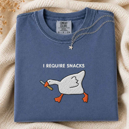 A folded slate blue crewneck tshirt with a white goose embroidery graphic holding a knife and the text 'I REQUIRE SNACKS'. A silver necklace with a star pendant is draped over the collar.