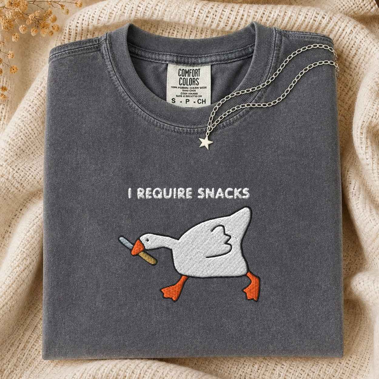 A folded dark gray t-shirt with an embroidery graphic of a white goose holding a knife in its beak, with the text 'I REQUIRE SNACKS' above it. A silver necklace with a star pendant is draped over the collar.