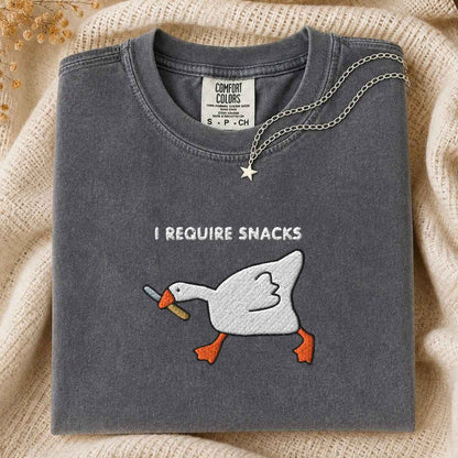 A folded dark gray t-shirt with an embroidery graphic of a white goose holding a knife in its beak, with the text 'I REQUIRE SNACKS' above it. A silver necklace with a star pendant is draped over the collar.