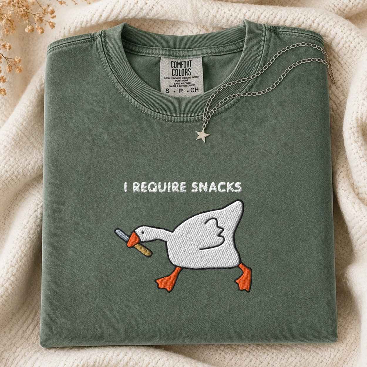 A folded sage green Comfort Colors t-shirt with a white goose graphic holding a knife and the text 'I REQUIRE SNACKS' on the front. The shirt is styled on a cream knit blanket with a silver star necklace draped over the collar.