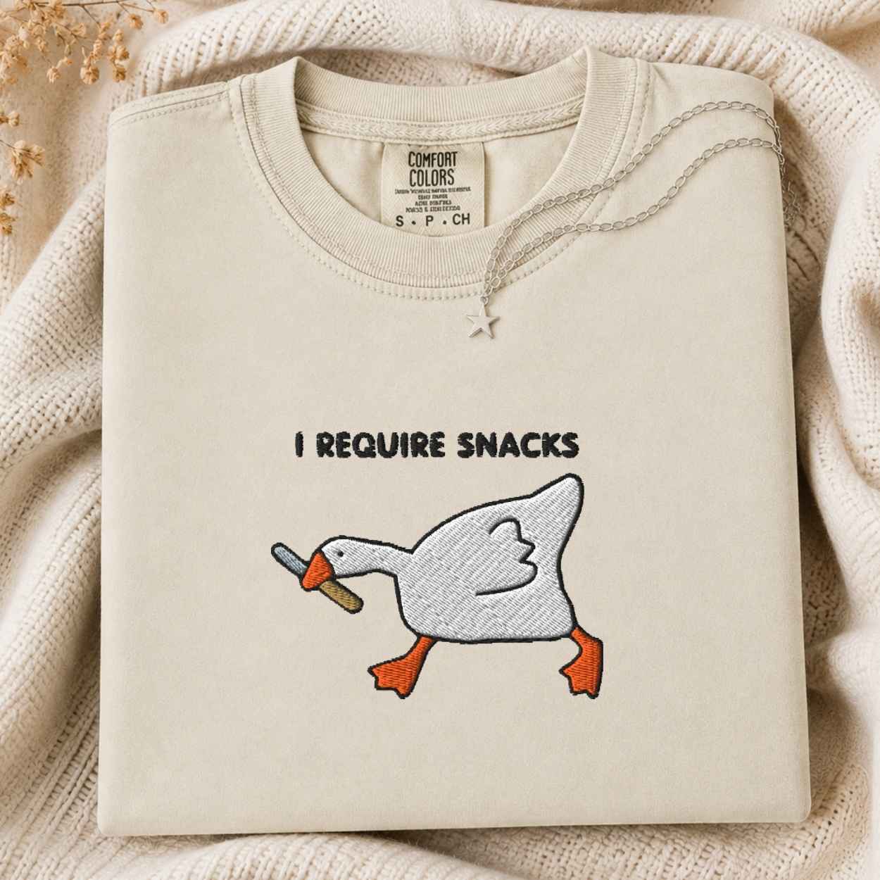 A folded cream-colored tshirt with an embroidery graphic of a white goose holding a knife, with the text 'I REQUIRE SNACKS' above it. The tshirt is laid flat on a cozy cream knit blanket with dried flowers in the corner.