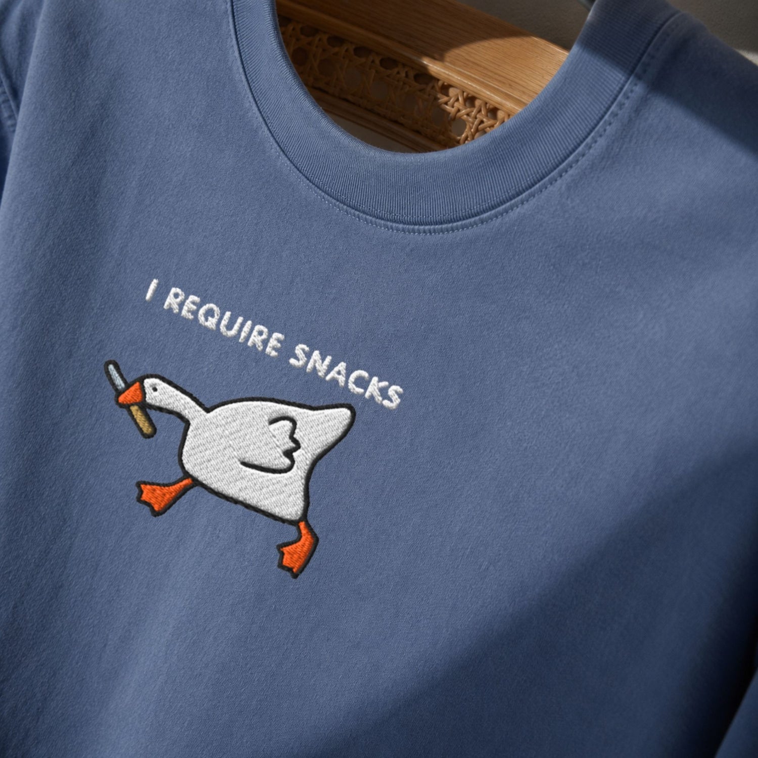 A close-up of a dusty blue crewneck t-shirt featuring an embroidered white goose holding a brown stick with the text 'I REQUIRE SNACKS' in white embroidery.