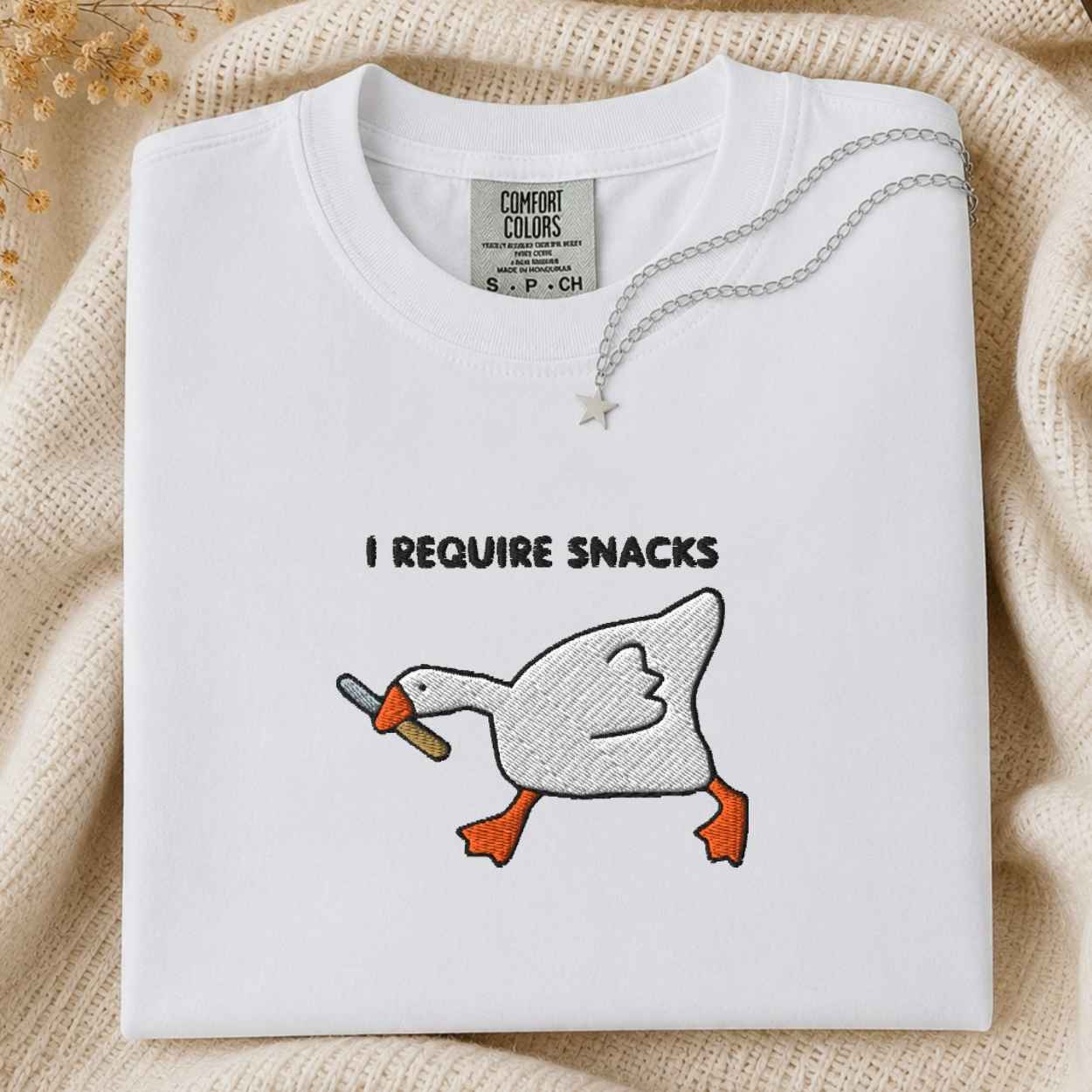 A folded white Comfort Colors t-shirt with a graphic of a goose holding a knife in its beak, with the text 'I REQUIRE SNACKS' above it. The shirt is styled on a beige knit blanket with a silver necklace draped over the collar.