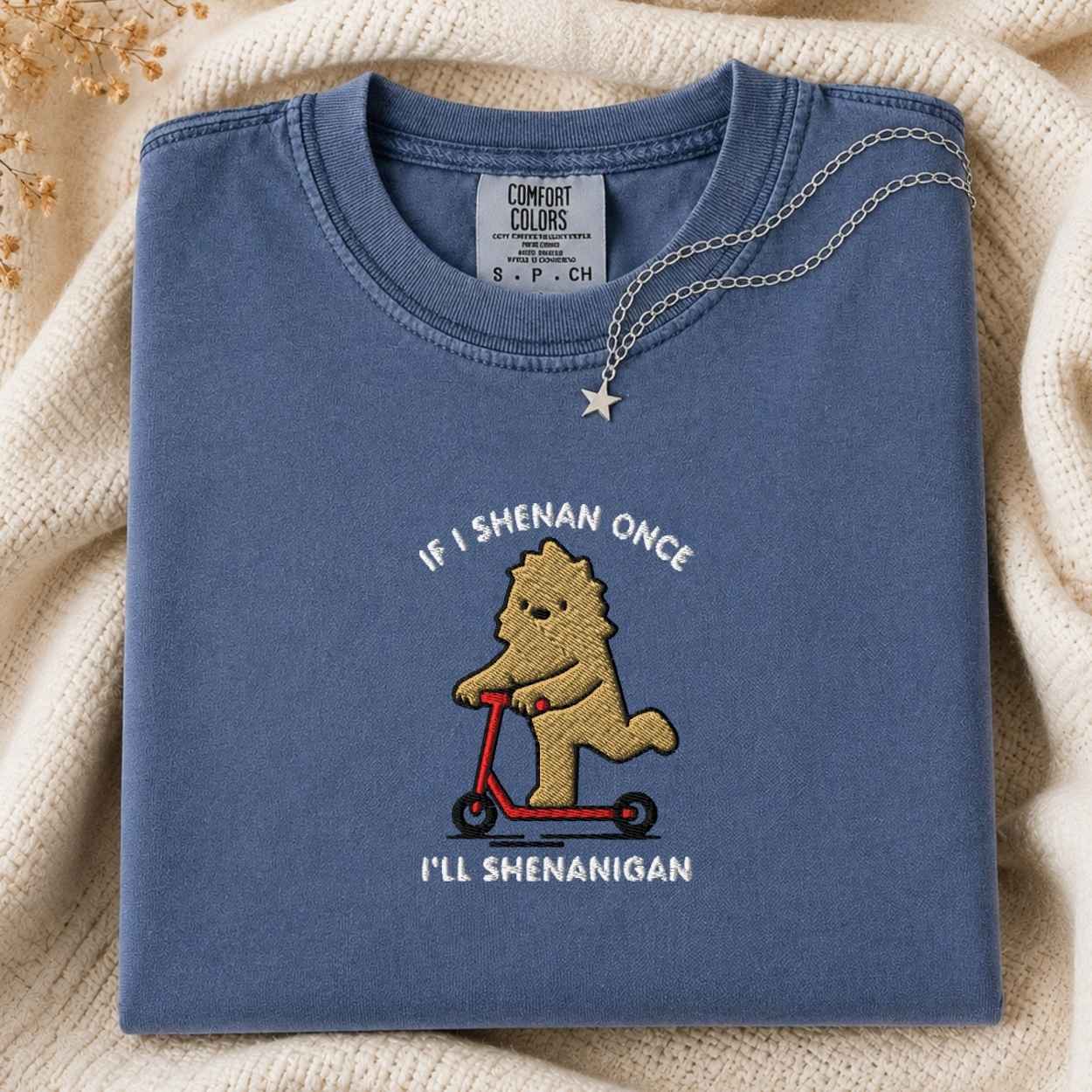 A folded slate blue crewneck t-shirt with a custom embroidered cartoon bigfoot riding a red scooter, displayed on a cream knit blanket with a silver necklace. The text is If I Shenan Once I'll Shenanigan