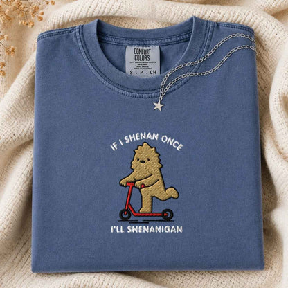 A folded slate blue crewneck t-shirt with a custom embroidered cartoon bigfoot riding a red scooter, displayed on a cream knit blanket with a silver necklace. The text is If I Shenan Once I'll Shenanigan