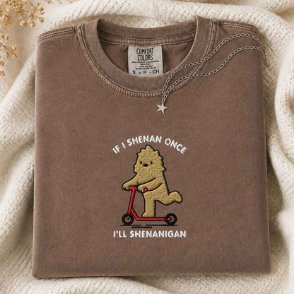 A folded brown crewneck tshirt with a graphic of a cartoon bigfoot riding a red scooter, displayed on a cream knit blanket with a small gold star necklace. The text is If I Shenan Once I'll Shenanigan