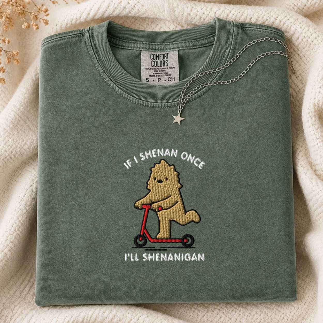 A folded sage green t-shirt with a cartoon bigfoot riding a red scooter embroidered on the front, the text is If I Shenan Once I'll Shenanigan styled on a cream knit blanket with a silver necklace.