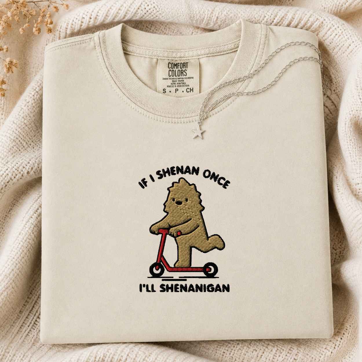 A folded cream-colored tshirt with a graphic of a brown cartoon bigfoot riding a red scooter, surrounded by a black text design saying If I Shenan Once I'll Shenanigan. The tshirt is displayed on a cozy, textured cream knit blanket with dried flowers in the corner.