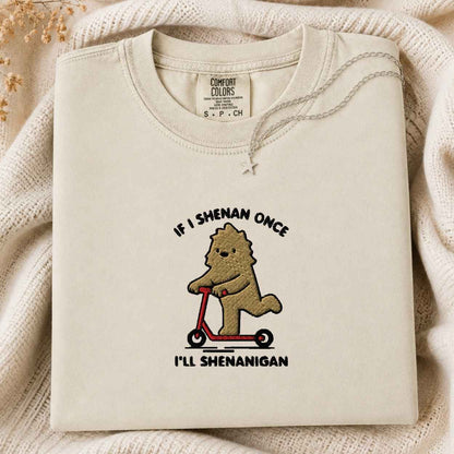 A folded cream-colored tshirt with a graphic of a brown cartoon bigfoot riding a red scooter, surrounded by a black text design saying If I Shenan Once I'll Shenanigan. The tshirt is displayed on a cozy, textured cream knit blanket with dried flowers in the corner.