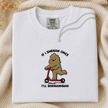 A folded white Comfort Colors t-shirt with a graphic of a bigfoot on a red scooter, displayed on a beige knit blanket with a silver necklace. The text is If I Shenan Once I'll Shenanigan