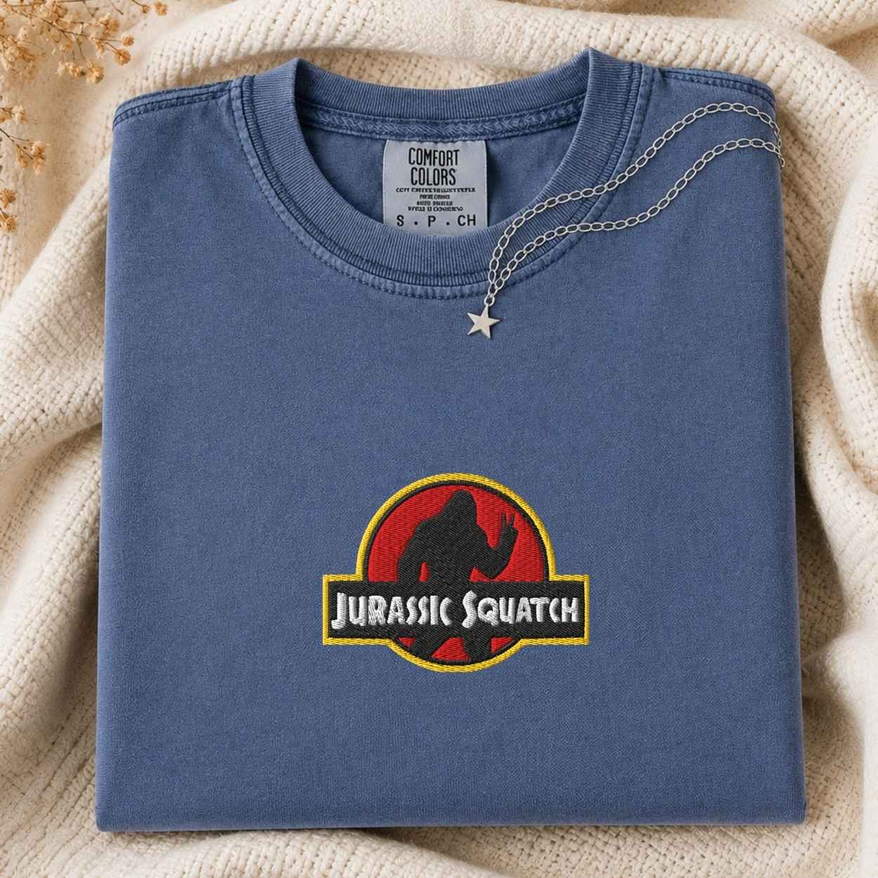 A folded blue t-shirt with a 'Jurassic Squatch' parody graphic on the front, styled on a cream knit blanket with a silver star necklace.